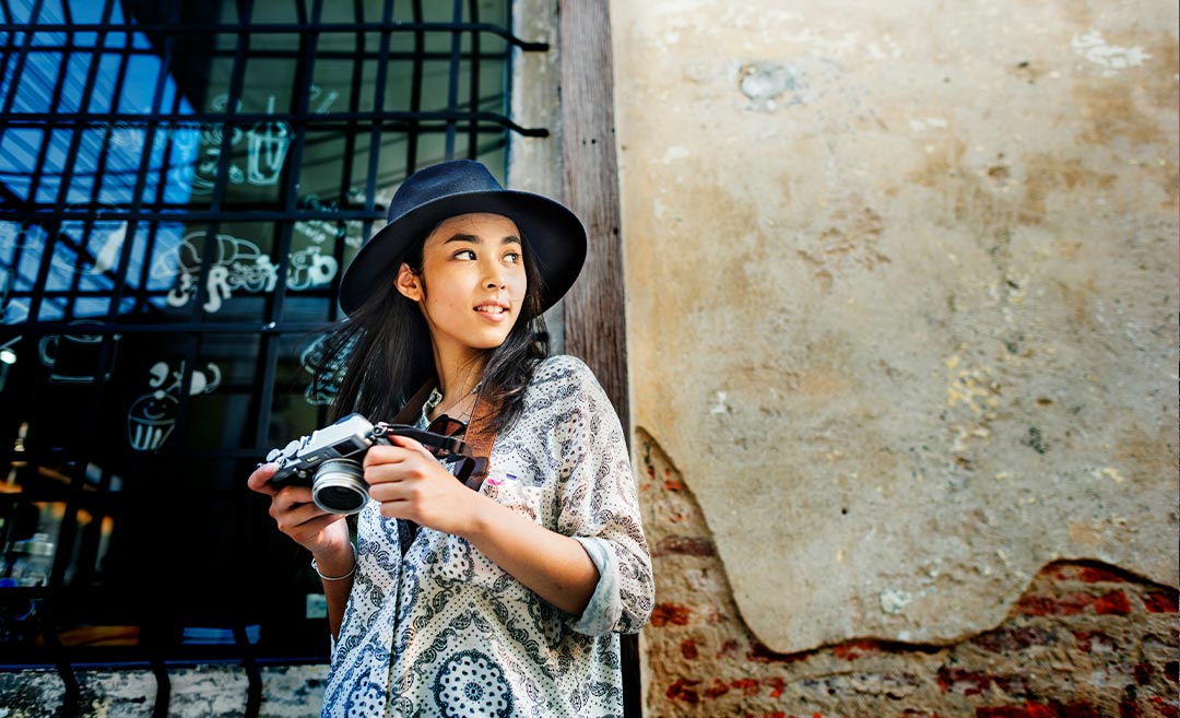 A woman with a camera and a hat stands before a building, illustrating intentional photography and beating travel fatigue.