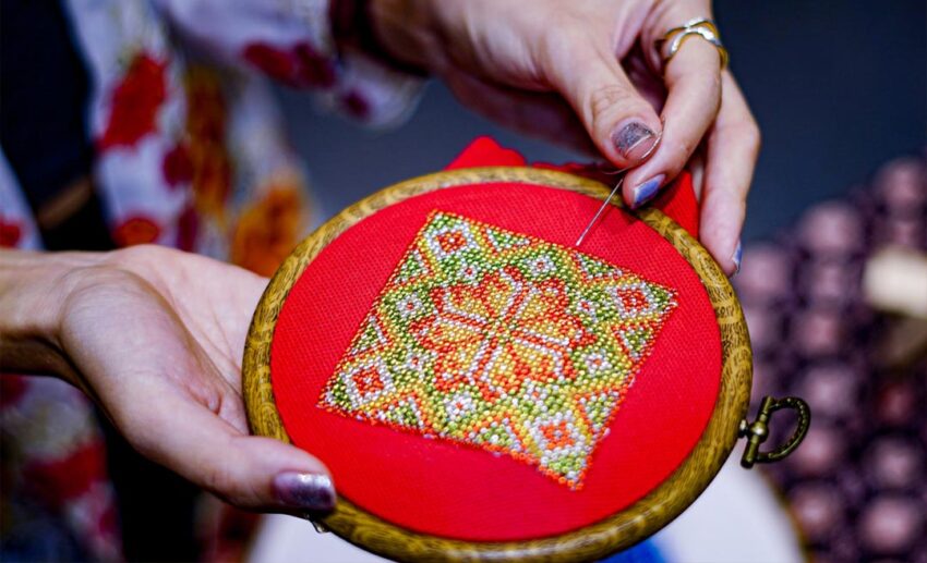  detailed close-up shot of Nyonya beadwork in progress, showing tiny coloured beads being meticulously sewn onto fabric for traditional Peranakan shoes.