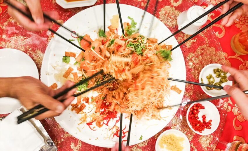 An overhead shot of a group of people using chopsticks to lift and toss Yee Sang ingredients high into the air during a prosperity toss (Lo Hei) in Malaysia.