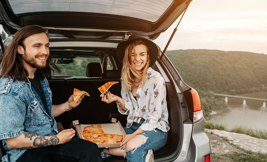 A young man and woman having fun sitting in a car trunk and eating pizza, travelling and road trip. 