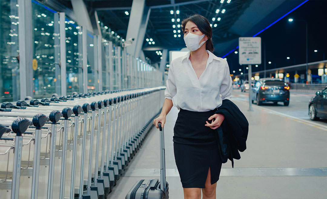 A woman in business attire wearing a face mask and pushing wheeled luggage outside a modern airport terminal.