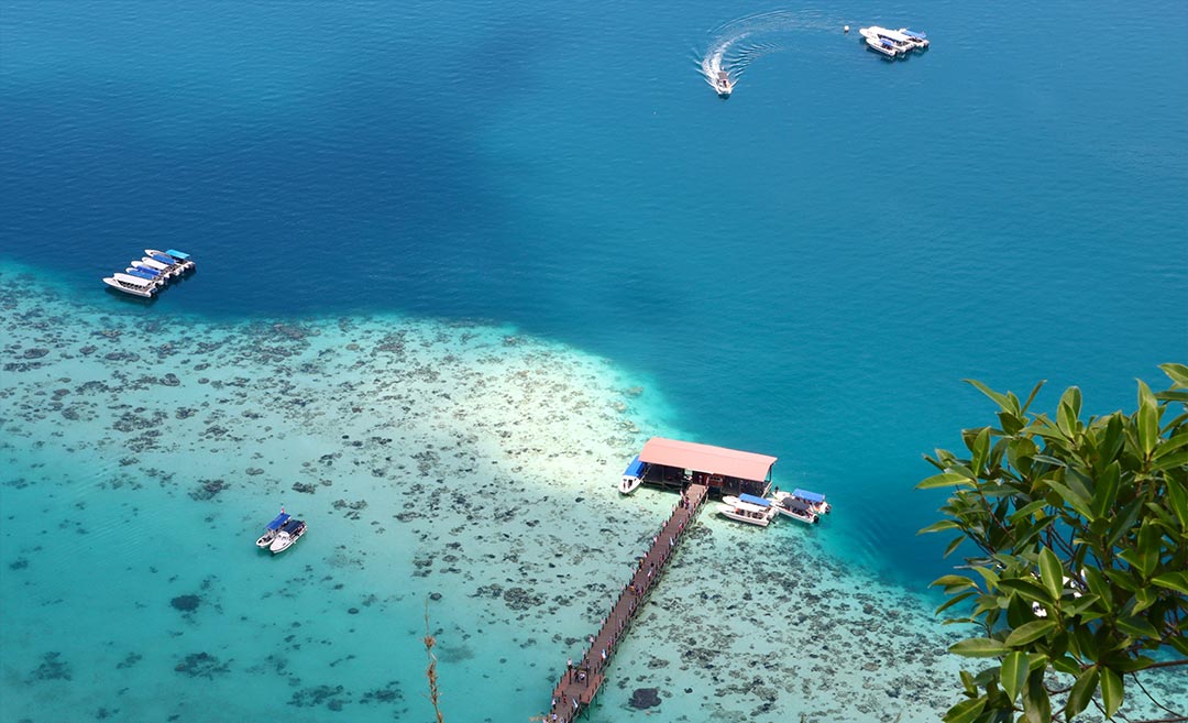 An aerial drone photograph of a wooden boat dock over clear turquoise water in Semporna, Sabah, a popular domestic travel destination in East Malaysia.