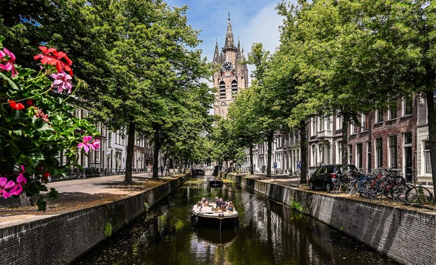 A small group of travellers enjoying a quiet boat cruise along a narrow Delft canal, with a historic clock tower in the distance and tree-lined streets filled with parked bicycles.