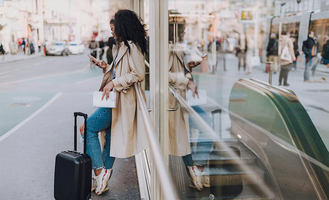A young woman with a travel trolley bag leaning against a glass subway entrance while checking messages on her smartphone.