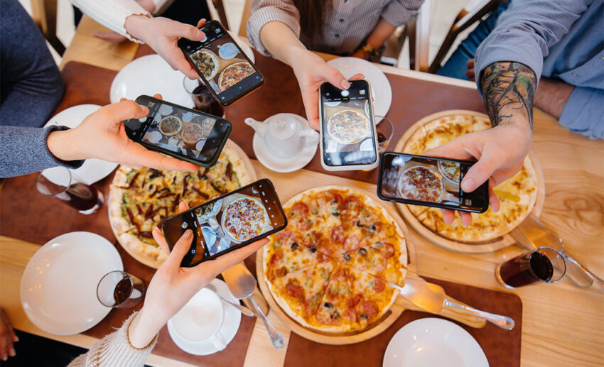 A group of friends holding smartphones above a dinner table, taking close-up photos of a delicious pizza before eating.