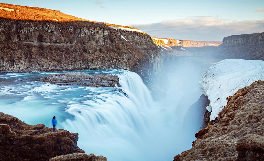 A traveller standing on a cliff overlooking the massive Gullfoss Falls in Iceland under a clear blue daytime sky.