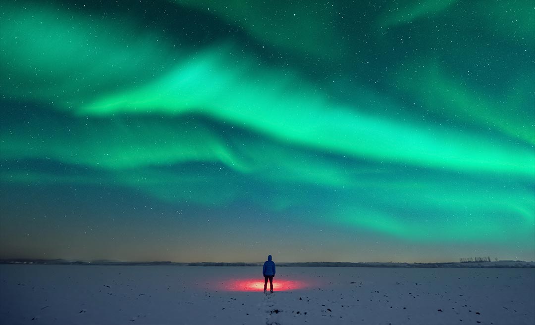 A traveller with a red flashlight standing on a snowy field under a vibrant starry sky and the Aurora Borealis northern lights.