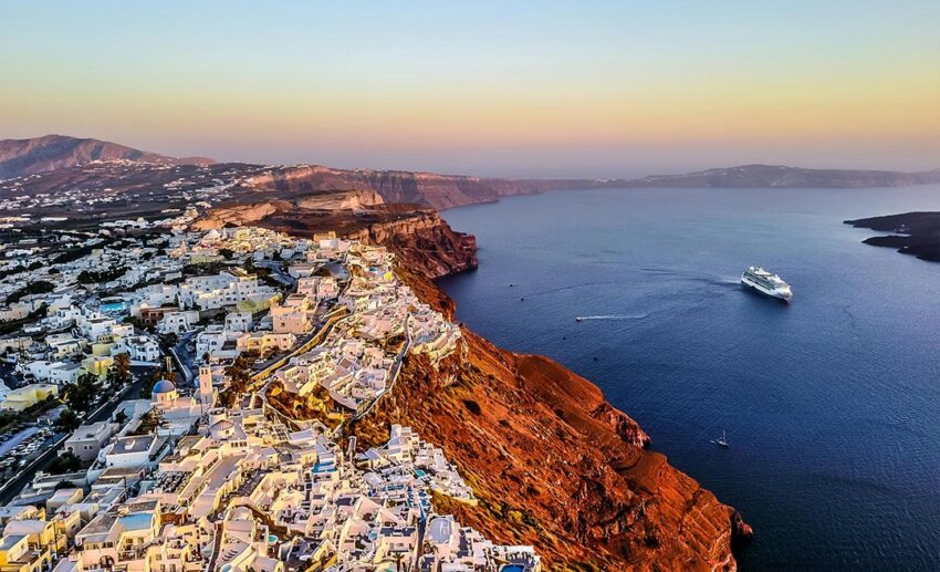 An aerial sunset view of the Santorini coastline in Greece, featuring a cruise ship on the Aegean Sea and iconic white-domed buildings.